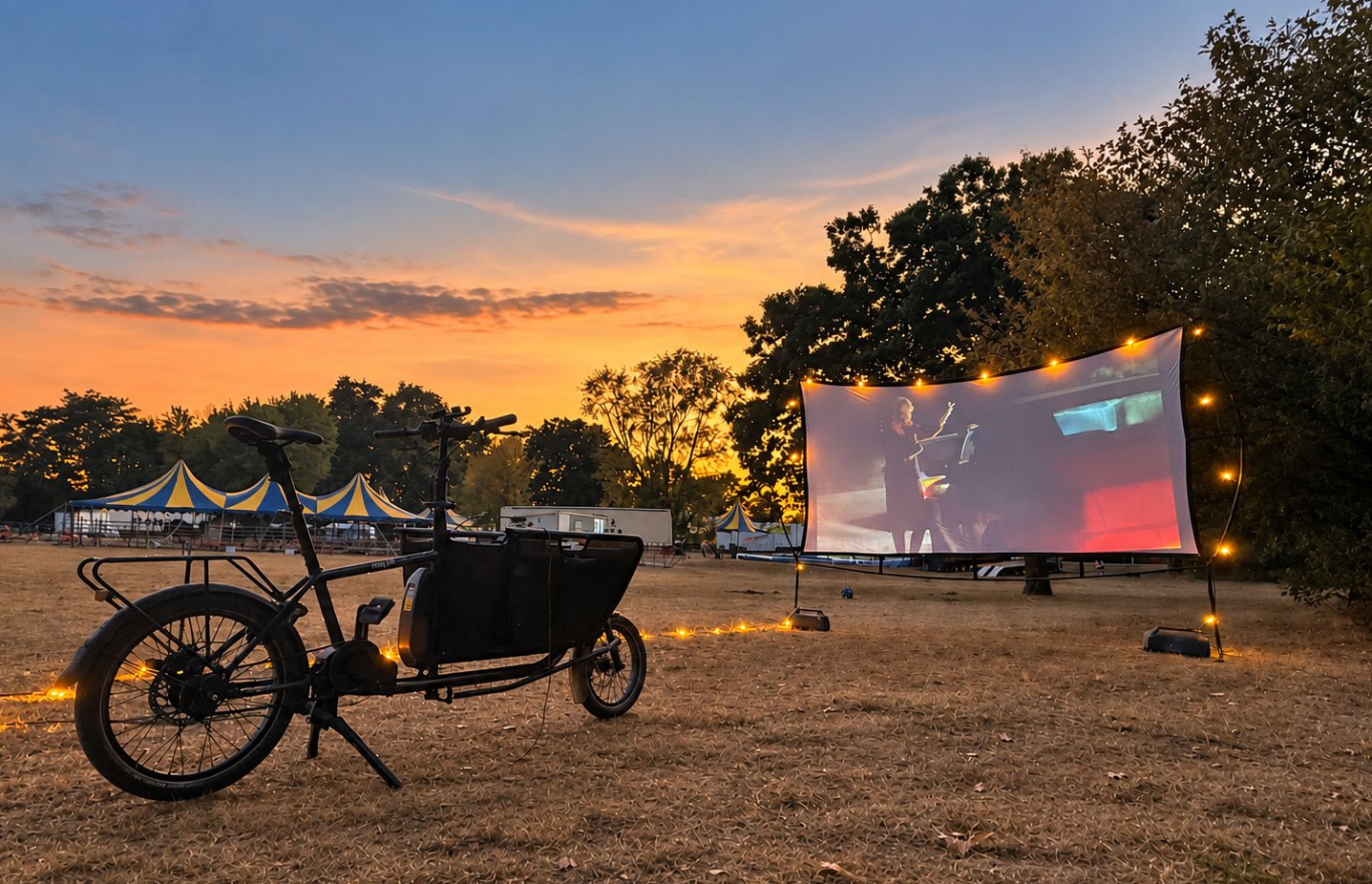 Lastenrad und Leinwand auf einer Wiese in der Abenddämmerung, im Hintergrund Zirkuszelte